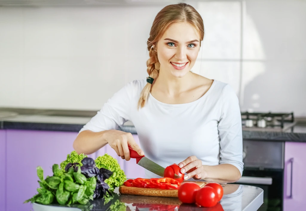Sarah Miller, author of Pure Plate Path, smiles while slicing a red tomato in a modern kitchen with fresh lettuce and basil nearby.