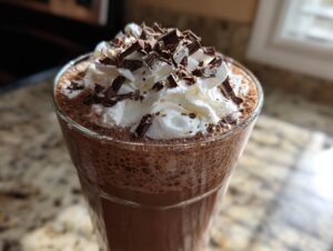Close-up of a tall glass filled with blended frozen hot chocolate, topped with whipped cream and chocolate shavings.