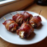 Five perfectly cooked bacon-wrapped dates stuffed with creamy cheese, served on a white plate.