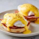 Two servings of classic eggs benedict featuring poached eggs, ham, and dripping hollandaise sauce on toasted English muffins.