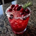 A vibrant red cranberry pomegranate margarita cocktail with a salted rim, topped with fresh cranberries and rosemary.