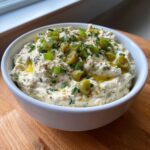 Close-up of a creamy dirty martini dip in a white bowl, garnished with sliced green olives, chives, and olive oil.