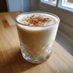 A glass of creamy eggnog topped with sprinkled cinnamon, sitting on a wooden table near a window.