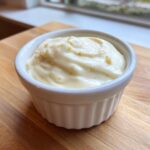 A close-up of creamy, pale yellow garlic sauce swirled in a small white ramekin on a wooden board.