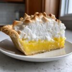 A perfect slice of lemon meringue pie showing bright yellow filling and tall, toasted meringue.
