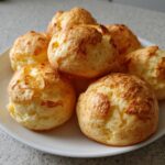 A close-up of several golden-brown, puffy brazilian cheese bread rolls stacked on a white plate.