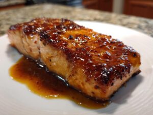 A perfectly cooked, thick fillet of salmon with a rich, shiny, dark glaze resting on a white plate.