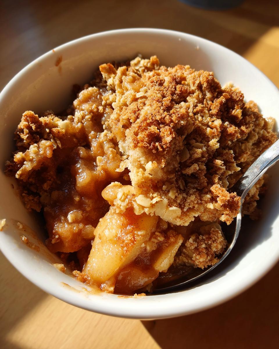 Close-up of a warm serving of apple crisp in a white bowl with a spoon, showing baked apples and crumb topping.