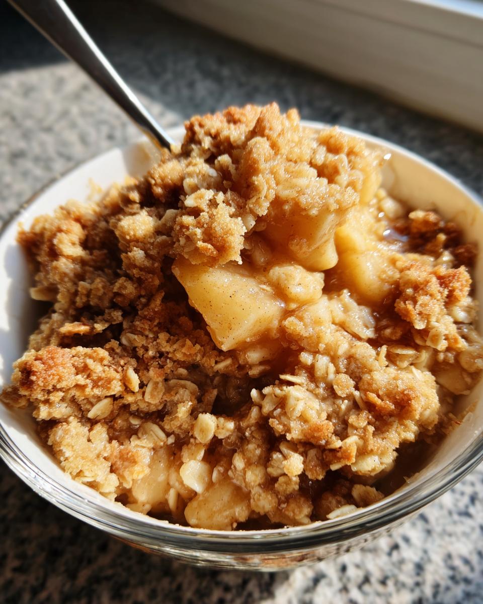 Close-up of a warm serving of apple crisp in a glass bowl, showing baked oat topping and soft spiced apples.