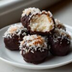 A stack of homemade almond joy truffles dipped in dark chocolate and topped with toasted coconut flakes.