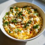 A close-up, appetizing view of a creamy corn dip topped with crumbled white cheese, fresh cilantro, and chili powder.