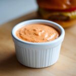 A close-up of creamy, orange-pink burger sauce speckled with spices, served in a white ramekin, with a blurred burger in the background.