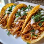 Close-up of three soft corn tortilla chicken tacos filled with shredded seasoned chicken, topped with diced white onion and fresh cilantro.