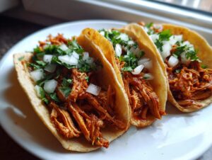 Three freshly made chicken tacos filled with shredded, saucy meat, topped with diced white onion and fresh cilantro.