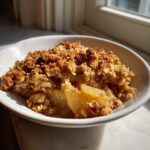 A close-up of a warm serving of apple crisp recipe in a white bowl, featuring soft cooked apples and a golden, crunchy oat topping.
