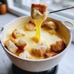 A piece of toasted bread is lifted from a white pot filled with smooth, melted cheese fondue, showing a long, gooey cheese pull.