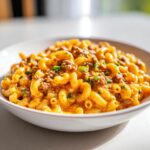 A close-up of a white bowl filled with cheesy taco pasta featuring elbow macaroni, seasoned ground meat, and green onions.