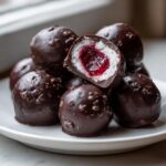A stack of homemade chocolate covered cherries, one cut open revealing the white filling and bright red cherry center.