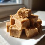A stack of creamy, light brown maple fudge cubes resting on a white square plate, illuminated by natural window light.