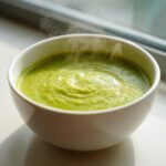 Close-up of a white bowl filled with hot, bright green creamy zucchini soup, showing steam rising.
