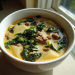 A close-up, appetizing shot of a bowl of Zuppa Toscana soup featuring creamy broth, chunks of potato, crumbled sausage, and wilted kale.