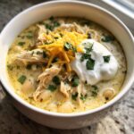 A close-up bowl of creamy crockpot white chicken chili topped with shredded chicken, white beans, cheese, sour cream, and cilantro.