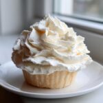 A single cupcake generously topped with swirls of light, fluffy vanilla buttercream frosting, sitting on a white plate.