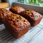 Four golden brown mini banana bread loaves topped with chocolate chips cooling on a wire rack.