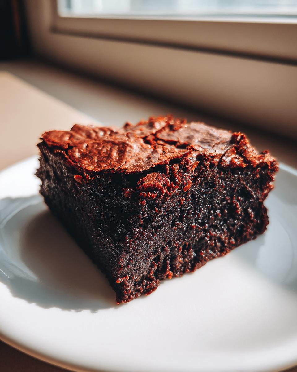 A rich, dark, fudgy square of date brownies sits on a white plate, highlighted by bright window sunlight.