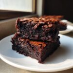 Two rich, fudgy date brownies stacked on a white plate, showing a shiny, crinkly top.