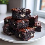 A stack of rich, fudgy Oreo brownies, showing chunks of white cream filling and topped with a mini Oreo cookie.