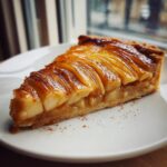 A perfect slice of golden, glazed apple tart featuring thinly sliced apples arranged on a flaky crust, sprinkled with cinnamon.