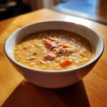 A close-up of a hearty bowl of Ham and bean soup, featuring chunks of ham and carrots, served on a wooden table.