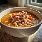 A close-up of a white bowl filled with hearty navy bean soup, topped with shredded meat and diced carrots.