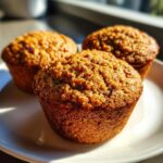 Close-up of three freshly baked honey bran muffins with textured tops sitting on a white plate in warm sunlight.