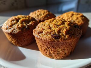 Close-up of four golden brown honey bran muffins with visible raisins, sitting on a white plate in bright sunlight.