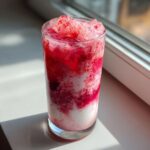 A tall glass showing layers of white milk and vibrant red strawberry syrup mixture, topped with crushed ice, perfect for strawberry milk.