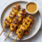 Four perfectly grilled chicken satay skewers served on a plate next to a small ramekin of peanut dipping sauce.
