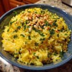 Close-up of bright yellow Mediterranean rice garnished with fresh parsley and toasted pine nuts in a blue bowl.