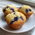 Close-up of three golden-brown blueberry protein muffins served on a small white plate.