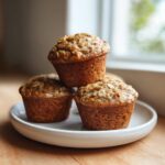 Three golden brown oatmeal muffins stacked on a small white plate, topped with visible rolled oats.