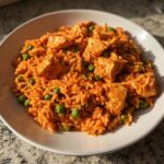 A close-up of a white bowl filled with vibrant orange Cajun chicken and rice mixed with bright green peas.