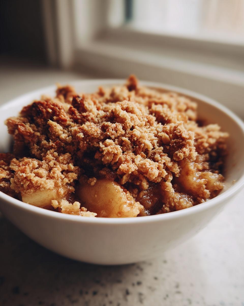A close-up shot of warm apple crumble with a thick, golden-brown topping served in a white bowl.
