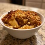Close-up of a single serving of warm apple crumble with a golden, crunchy topping in a white bowl.