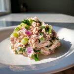 A mound of creamy salmon salad mixed with chopped red onion, celery, and herbs, served on a white plate.