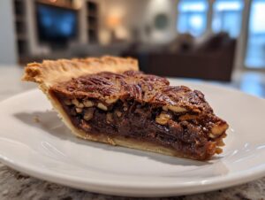 A close-up of a generous slice of chocolate bourbon pecan pie showing the flaky crust and rich, dark filling topped with pecans.