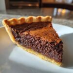 Close-up of a rich, fudgy slice of chocolate chess pie with a golden crust, sitting on a white plate.