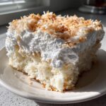 A close-up slice of moist coconut poke cake topped with thick white frosting and toasted coconut flakes.
