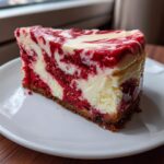 A close-up of a decadent slice of red velvet cheesecake featuring rich red cake layers swirled with creamy white cheesecake filling.
