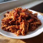 A mound of saucy, shredded Slow Cooker Pulled Pork piled high on a white plate, catching bright sunlight.